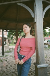 a young woman in a pink off the shoulder top and jeans leaning against a gazebo